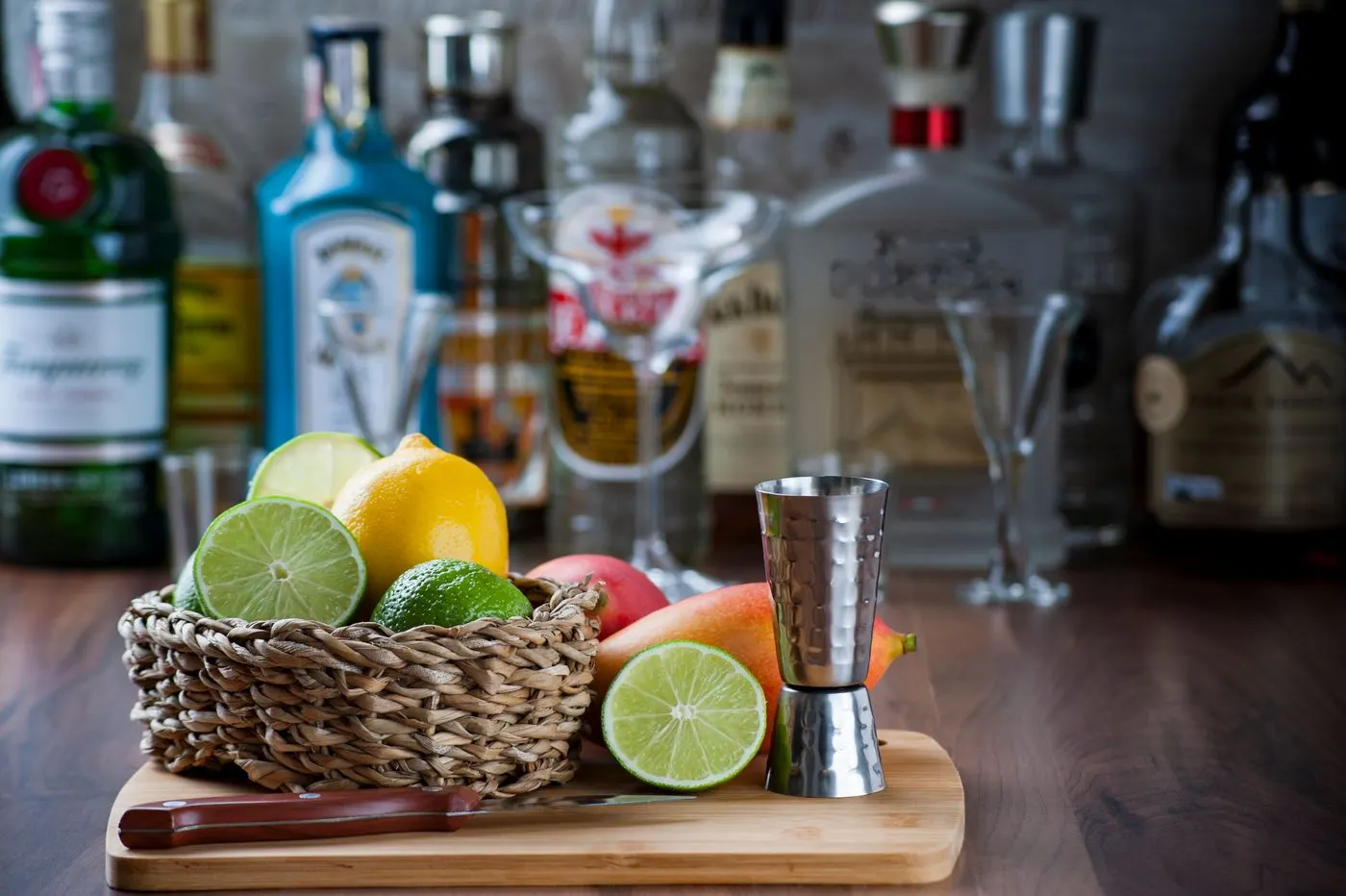 A jigger, fresh citrus in a basket and spirit bottles on a wooden bar top, the basics of a home bar