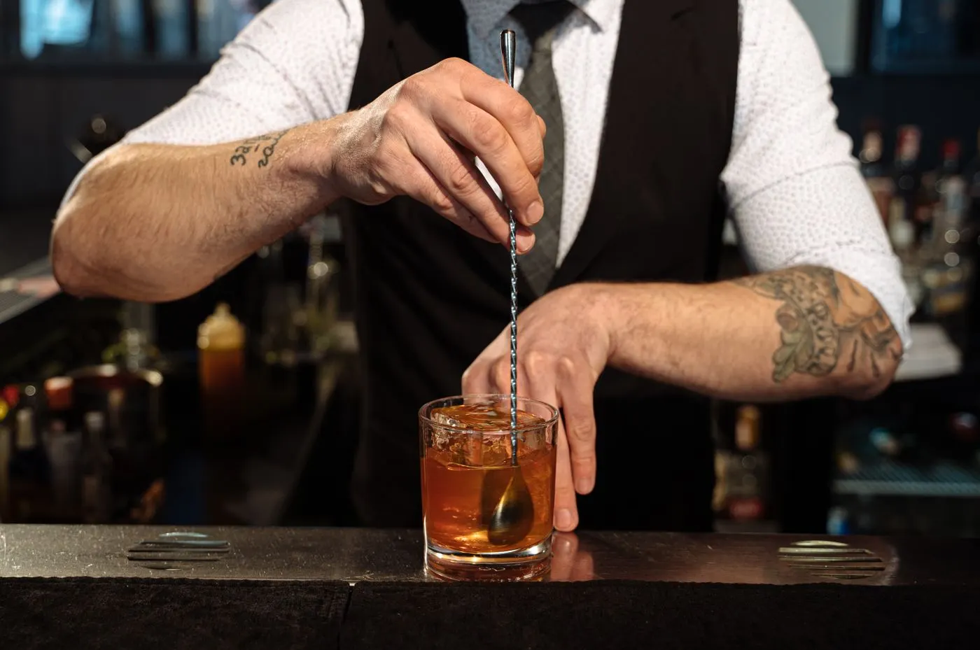A bartender stirring an amber cocktail with a long bar spoon in a rocks glass