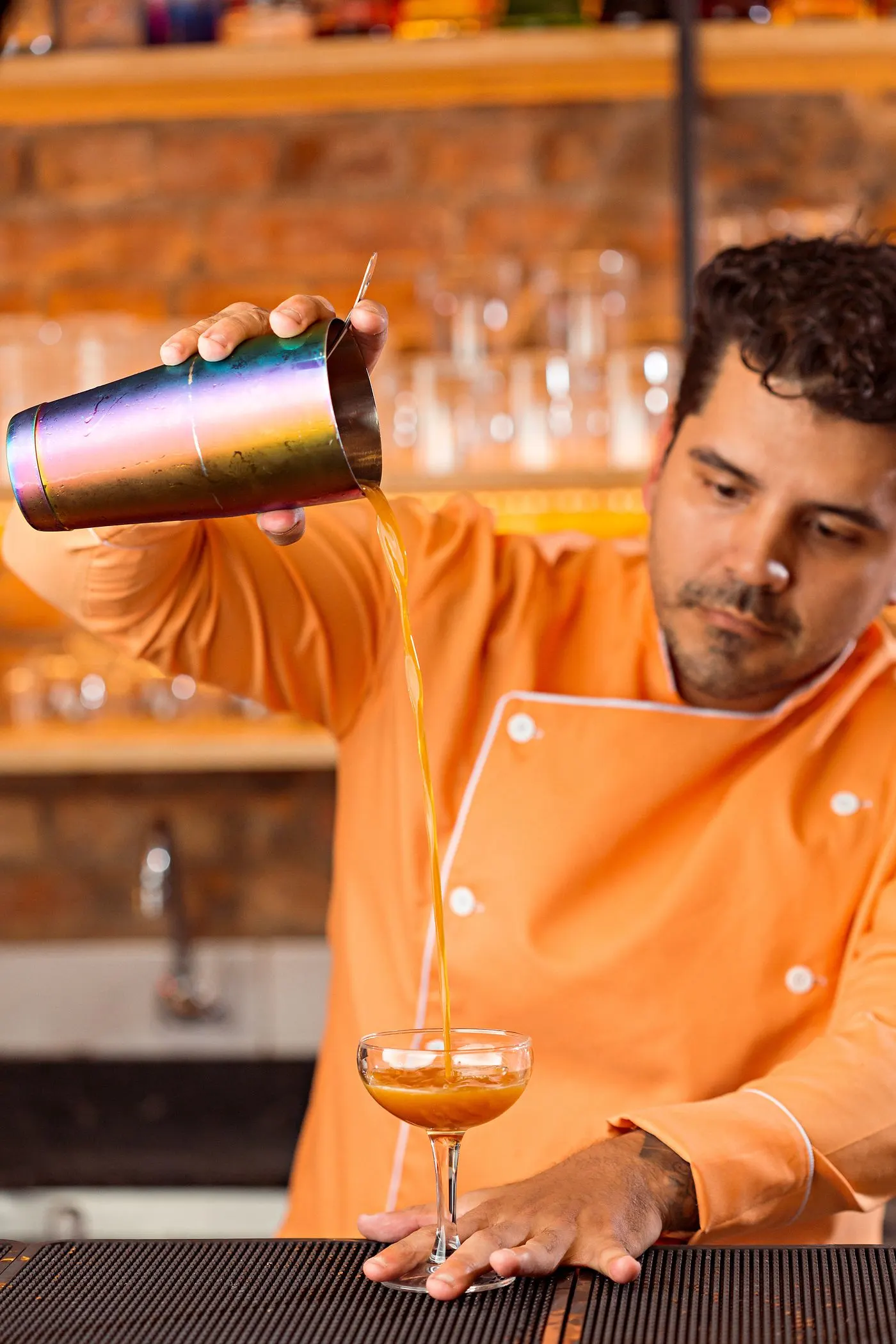 A bartender pouring a shaken cocktail from a metal shaker into a coupe glass