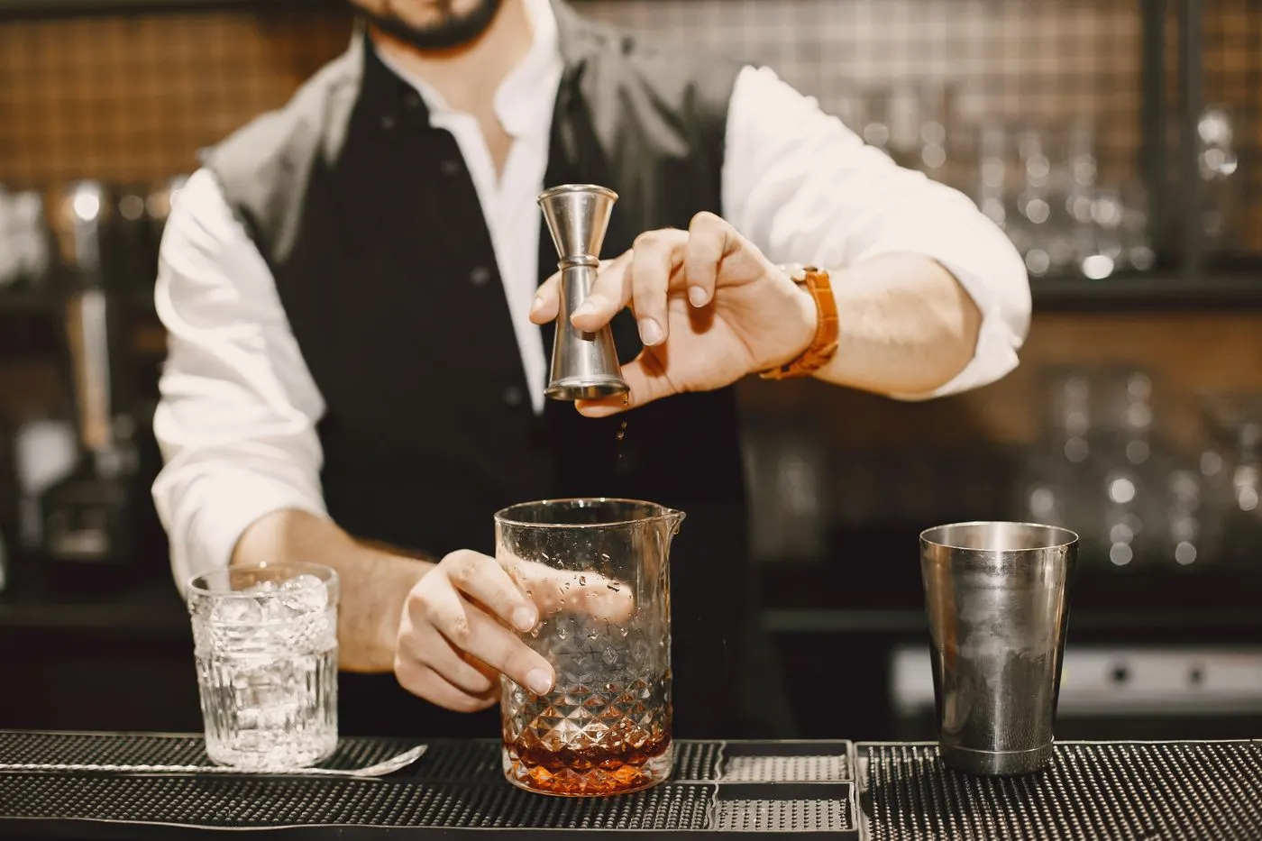 A bartender measuring a spirit with a jigger over a mixing glass, with a shaker tin beside it