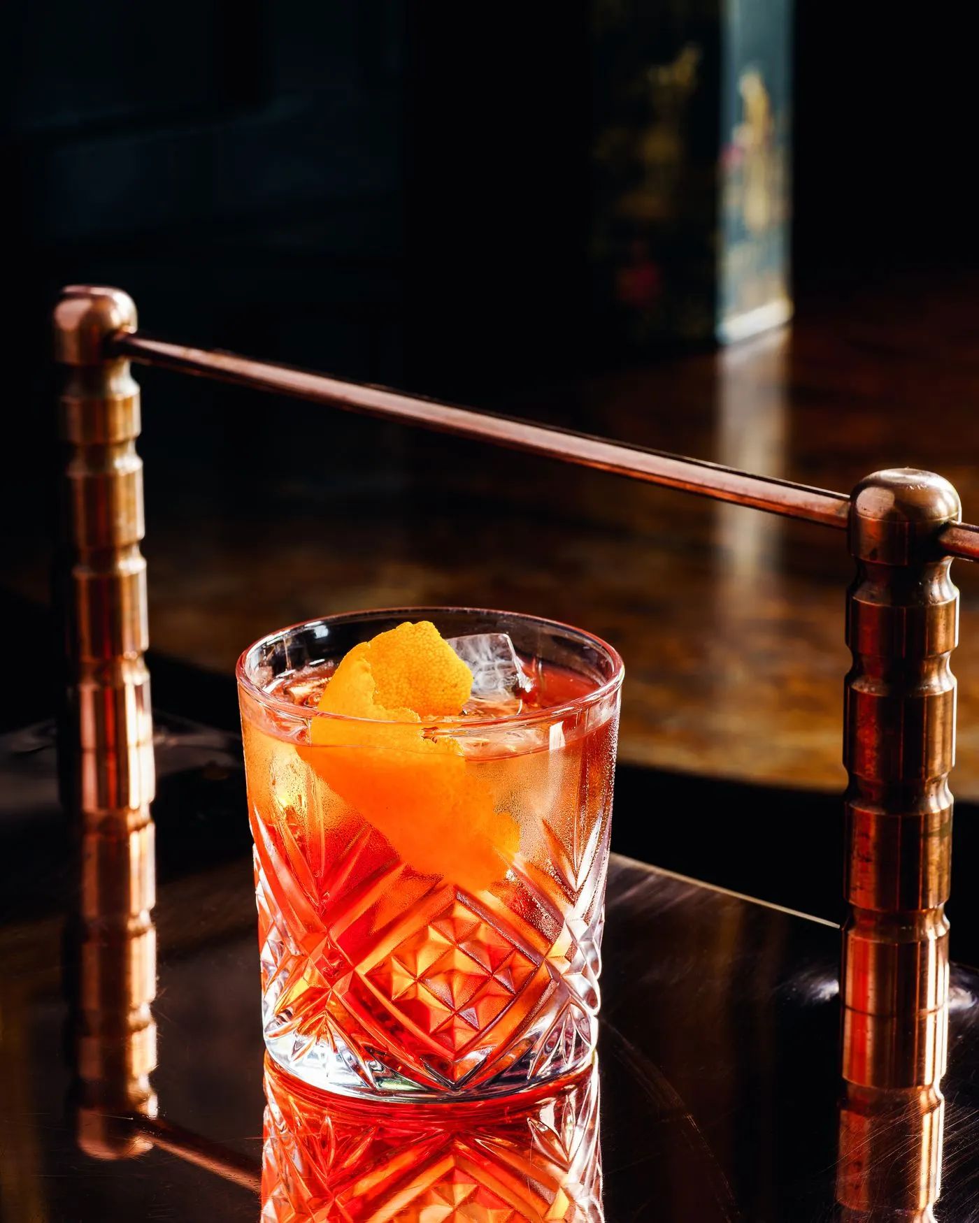 A Negroni in a faceted glass with an orange peel, standing on a copper rail in a dim bar
